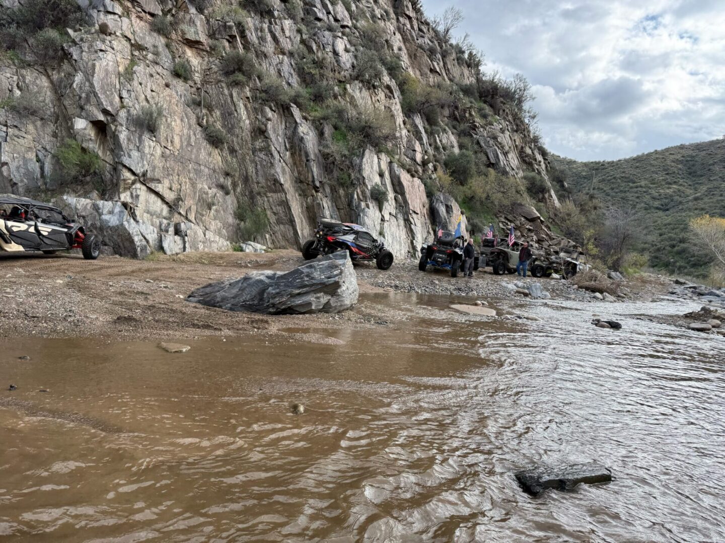 UTVs parked beside rocky cliff near muddy river