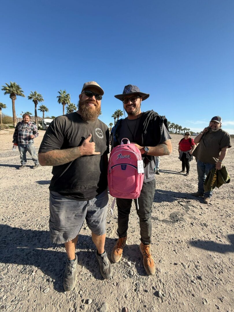 Two men outdoors holding pink backpack, palm trees