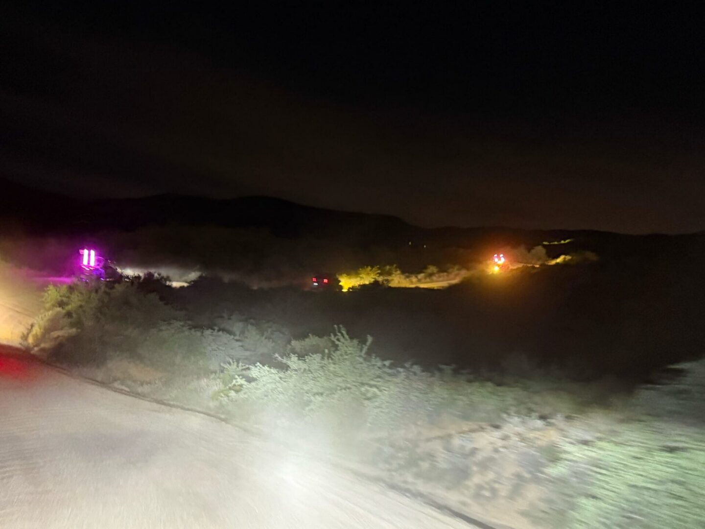 Headlights illuminating desert road with distant colored lights