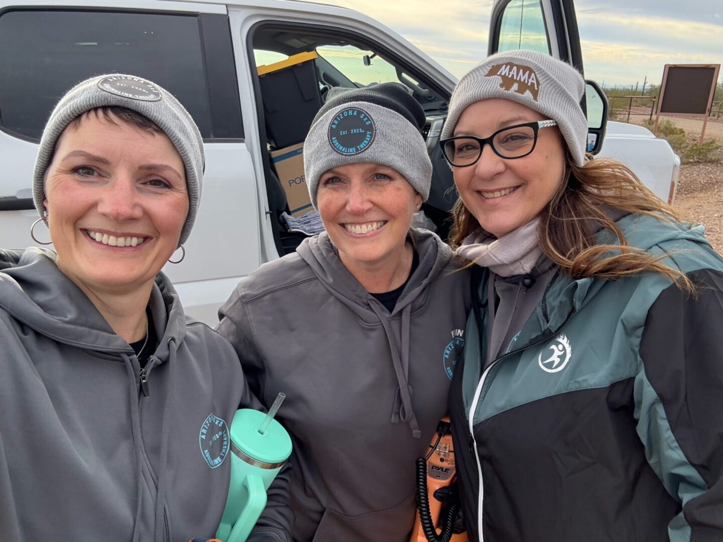 Three women in beanies smiling beside white vehicle